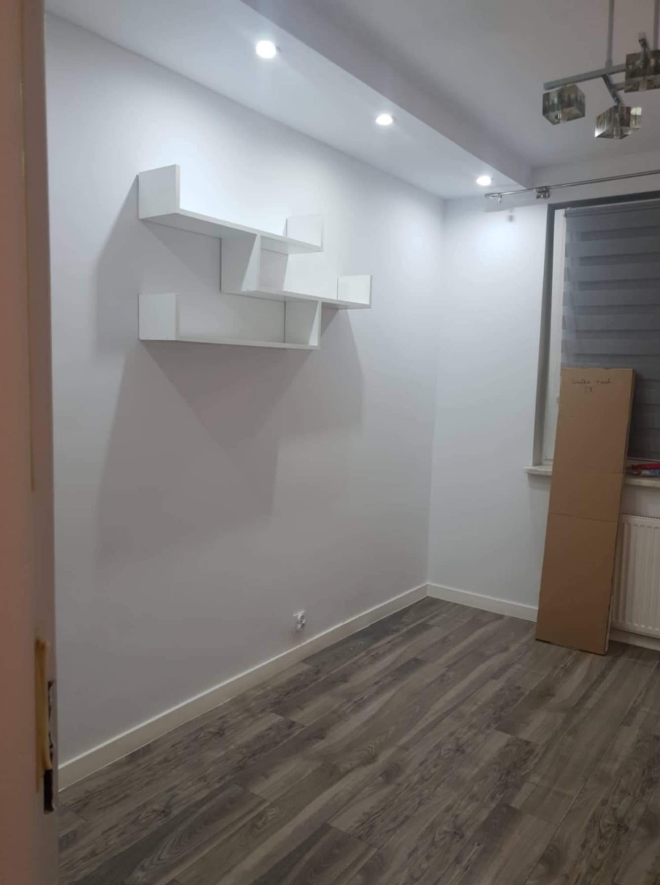 Pusty pokój po remoncie z szarą podłogą imitującą drewno, białe ściany z zamontowanymi białymi półkami na książki o nieregularnym kształcie, karton oparty o ścianę przy oknie.