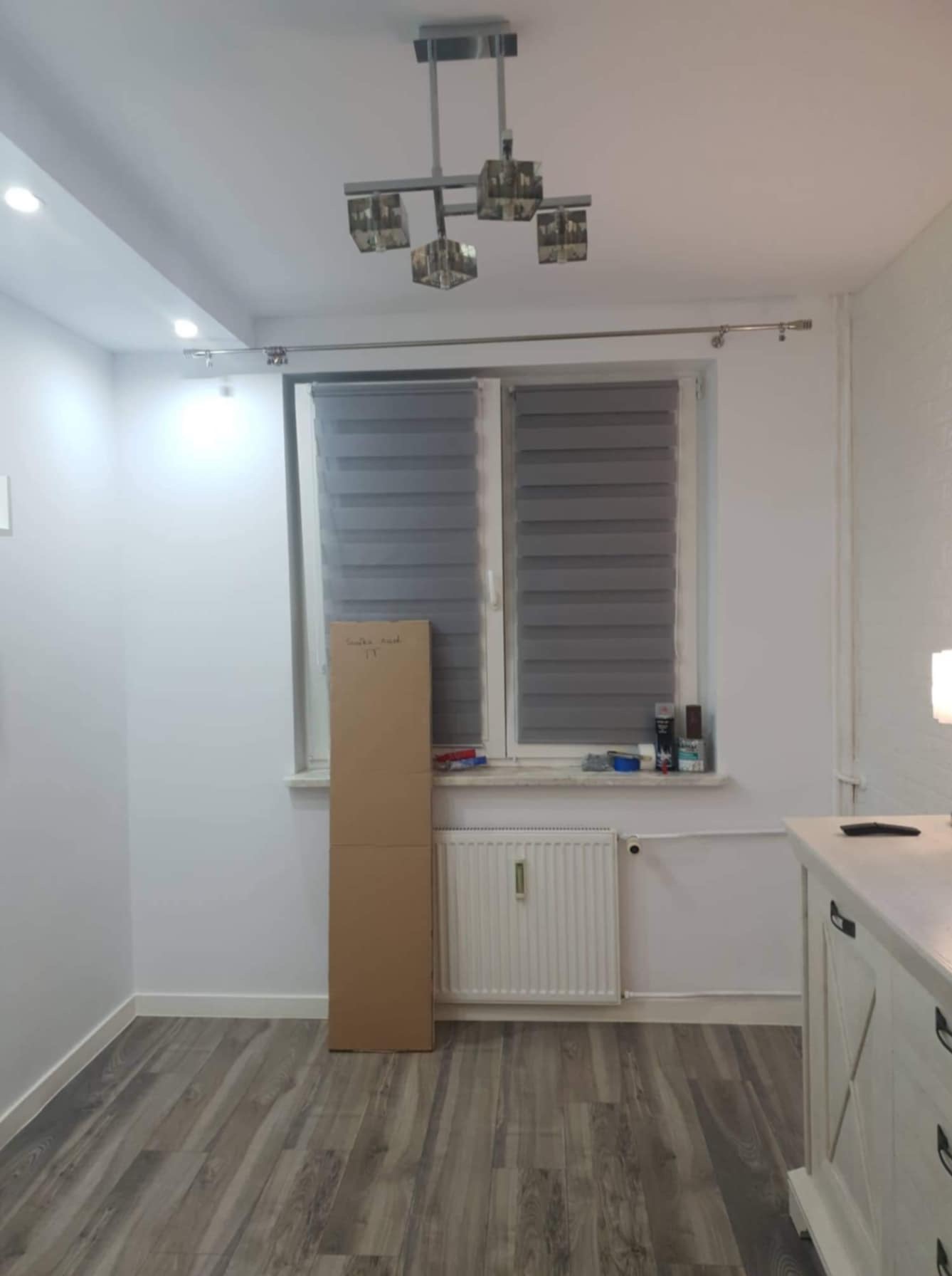 Pusta biała ściana w pokoju z szarymi roletami w oknach, kartonowe pudło opierające się o ścianę, grzejnik pod oknem, drewnopodobna podłoga, widoczna część białej komody z szufladami, nowoczesna...