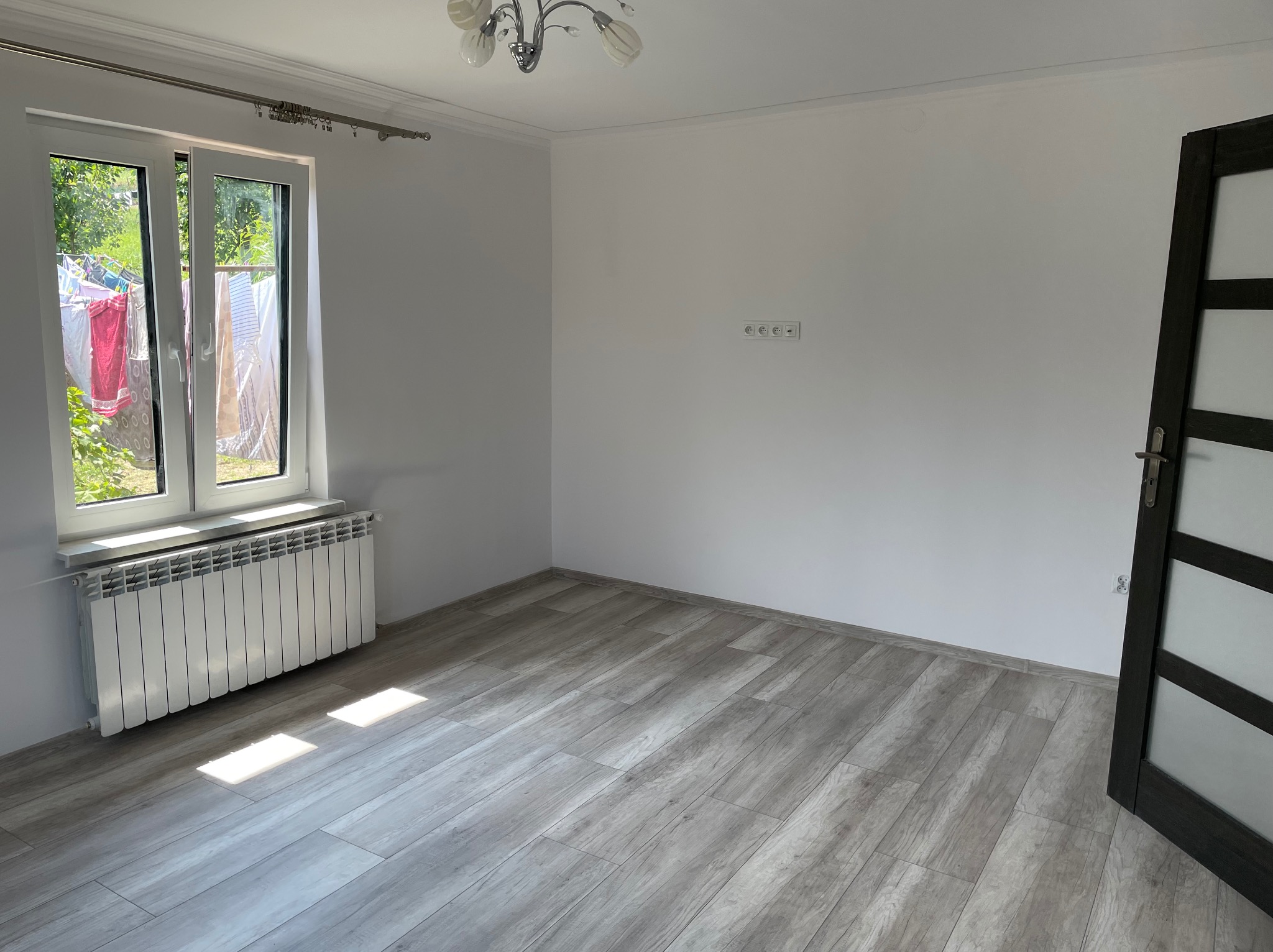 Puste, odnowione pomieszczenie z szarymi panelami podłogowymi, białymi ścianami, grzejnikiem pod oknem i czarnymi drzwiami z przeszkleniami, widok na suszące się pranie za oknem.