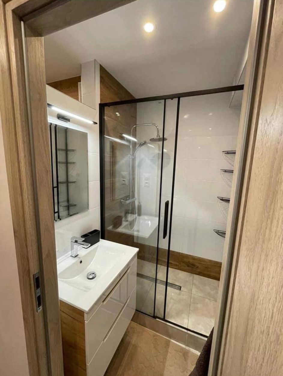 Nowoczesna łazienka z kabiną prysznicową typu walk-in z czarnymi profilami, białą umywalką nablatową z drewnianą szafką i lustrem z oświetleniem LED.