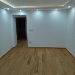 NOWYKONT44 Kompleksowe Usługi Remontowo - Wykończeniowe Marcin Bielatowicz - Pusty pokój po remoncie z jasnymi panelami podłogowymi, białymi ścianami, sufitem podwieszanym z oświetleniem punktowym i widocznym gniazdkiem elektrycznym.