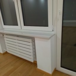 NOWYKONT44 Kompleksowe Usługi Remontowo - Wykończeniowe Marcin Bielatowicz - Biały grzejnik panelowy zamontowany pod oknem z parapetem, na drewnianej podłodze, widok z boku.