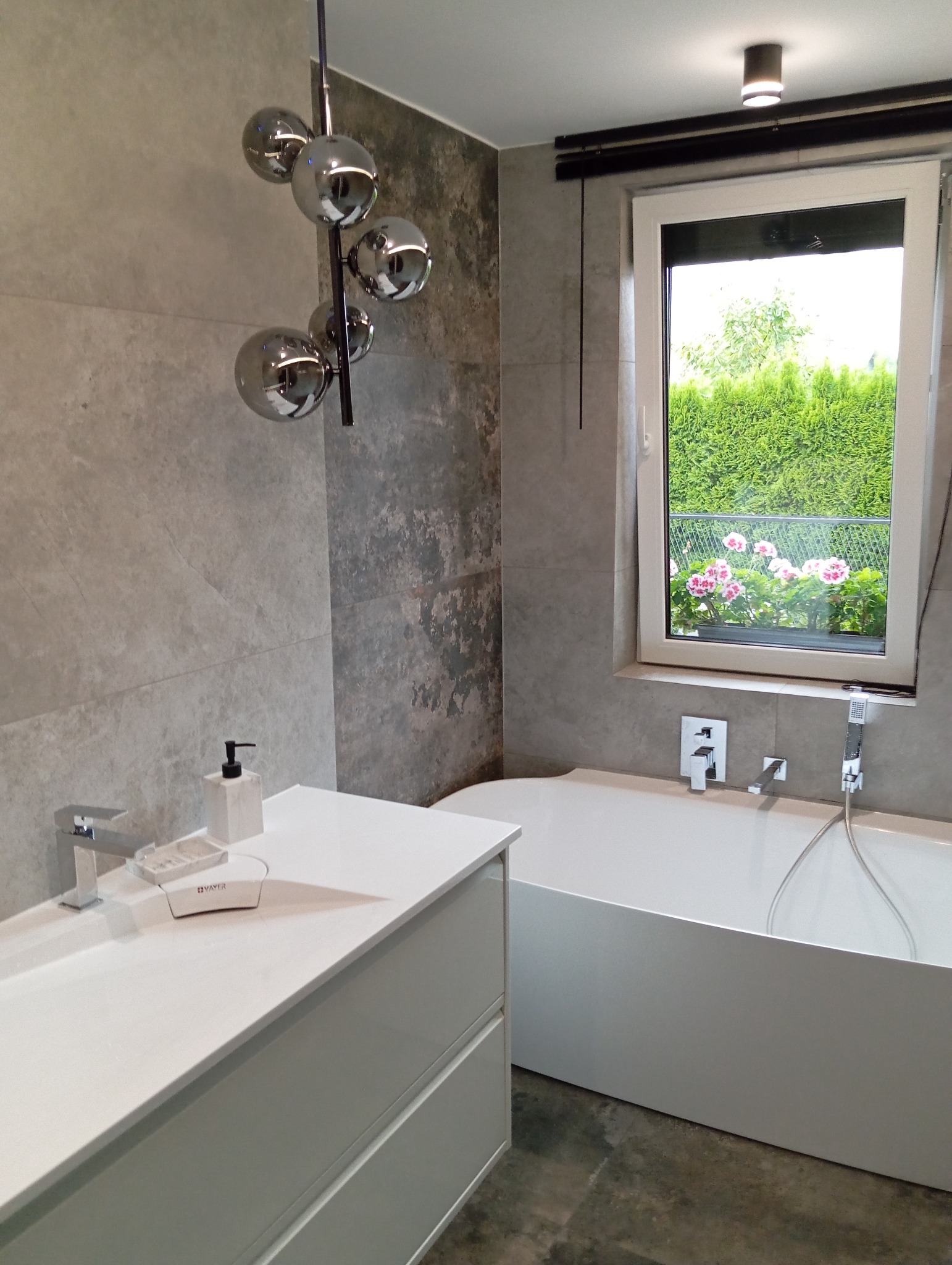 Nowoczesna łazienka z szarą płytką imitującą kamień, białą umywalką z szafką i wanną, oświetlona lampą kulistą i światłem wpadającym przez okno z widokiem na ogród.