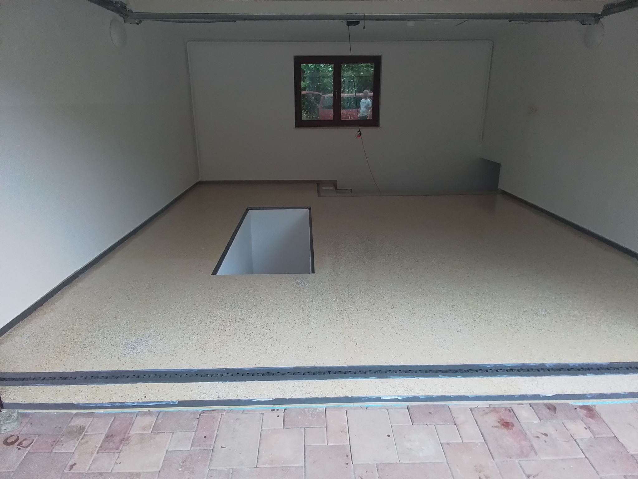 Wykończone wnętrze pomieszczenia z beżową, nakrapianą posadzką żywiczną, widoczne otwory w podłodze, okno z odbiciem osoby, białe ściany i metalowe elementy konstrukcyjne pod sufitem.