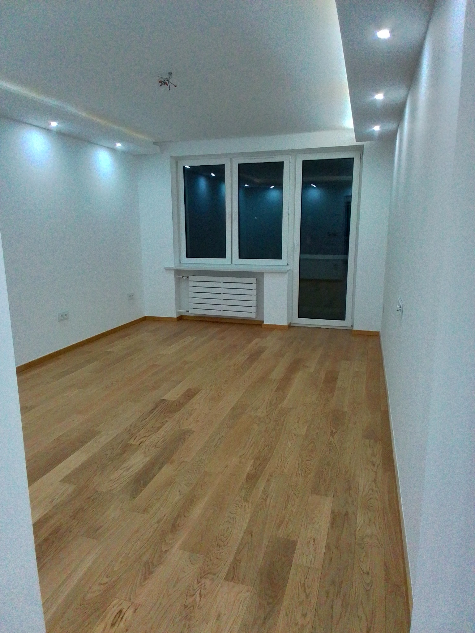 Pusty pokój po remoncie z drewnianą podłogą, białymi ścianami, oświetleniem sufitowym LED i oknem z grzejnikiem.