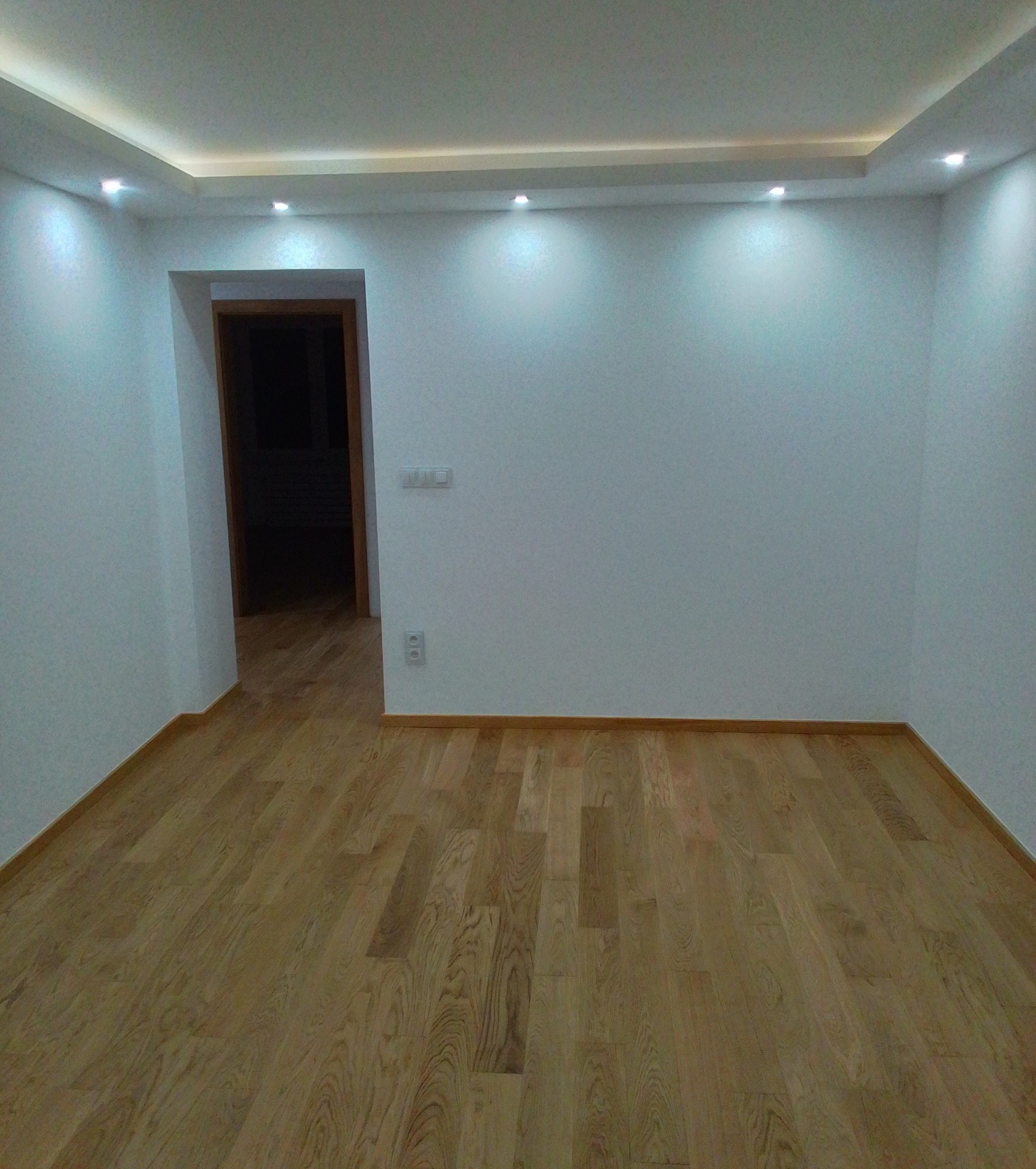 Pusty pokój po remoncie z jasnymi panelami podłogowymi, białymi ścianami, sufitem podwieszanym z oświetleniem punktowym i widocznym gniazdkiem elektrycznym.