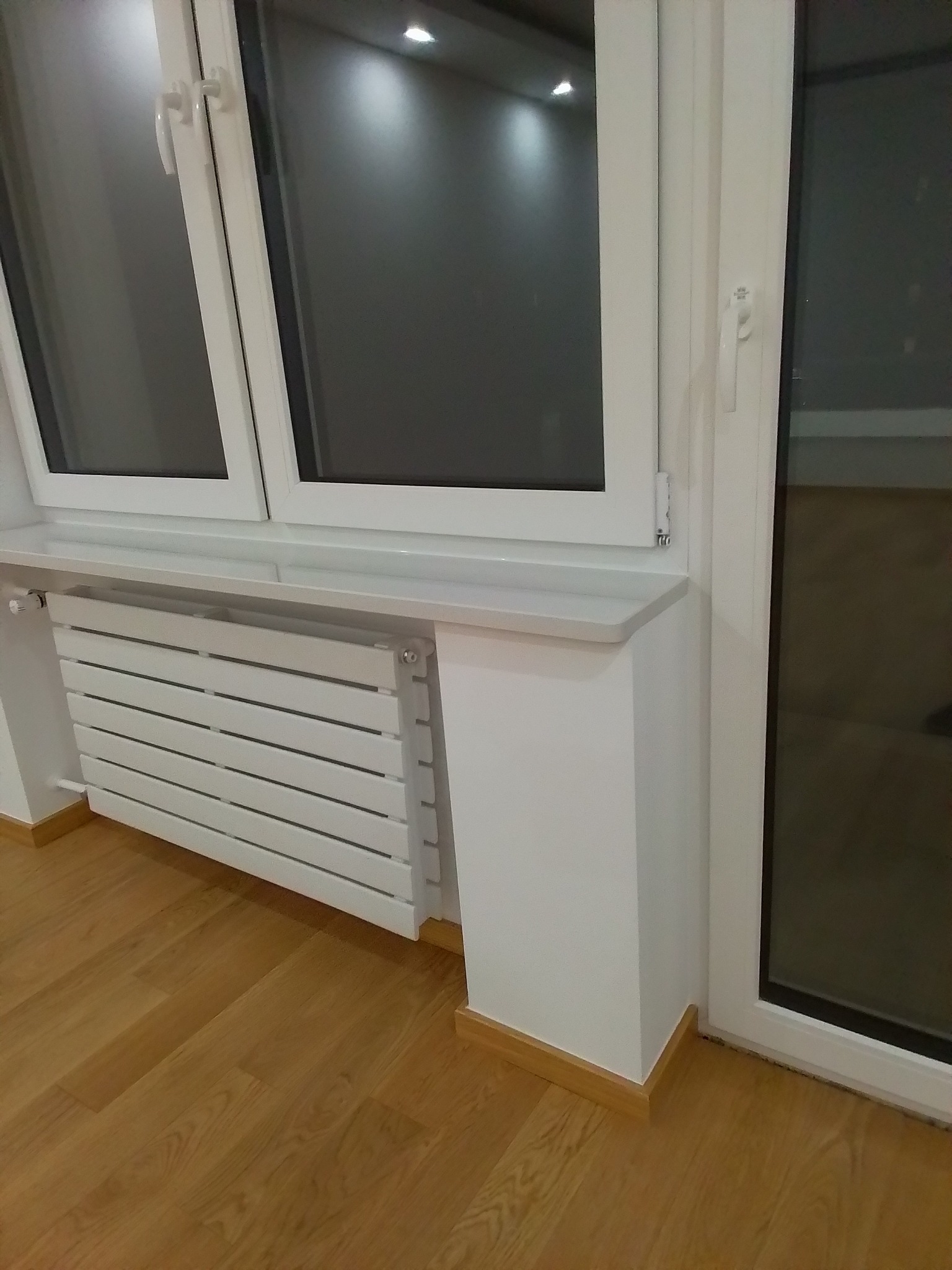 Biały grzejnik panelowy zamontowany pod oknem z parapetem, na drewnianej podłodze, widok z boku.