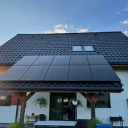 Czarny panel słoneczny zamontowany nad tarasem domu jednorodzinnego z ciemną dachówką, widoczne rośliny w donicach i drabina oparta o ścianę