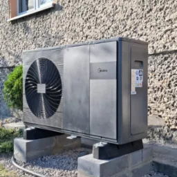 Szara pompa ciepła Midea zamontowana na betonowych blokach fundamentowych przed budynkiem z oknem.