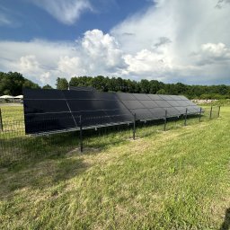 PROZE Jakub Kożuszko - Rząd paneli fotowoltaicznych zabezpieczony metalową siatką na trawiastym terenie, z widocznym lasem w tle i pochmurnym niebem.