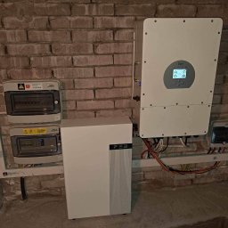 PROZE Jakub Kożuszko - Instalacja wewnętrzna: biały magazyn energii FEB, inwerter Deye z ekranem i skrzynki elektryczne na tle ceglanej ściany w piwnicy.