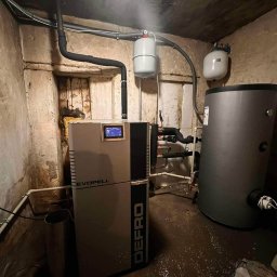 PROZE Jakub Kożuszko - Kotłownia z kotłem na pellet marki Defro Evopell i zbiornikiem akumulacyjnym w pomieszczeniu o surowych, betonowych ścianach. Widoczne rury i osprzęt instalacyjny.