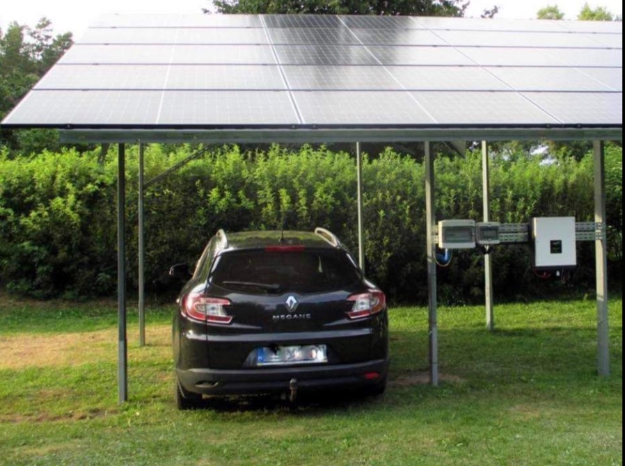 Czarny samochód Renault Megane zaparkowany pod wiatą z paneli słonecznych, z widocznymi inwerterami i zielonym tłem z krzewów.