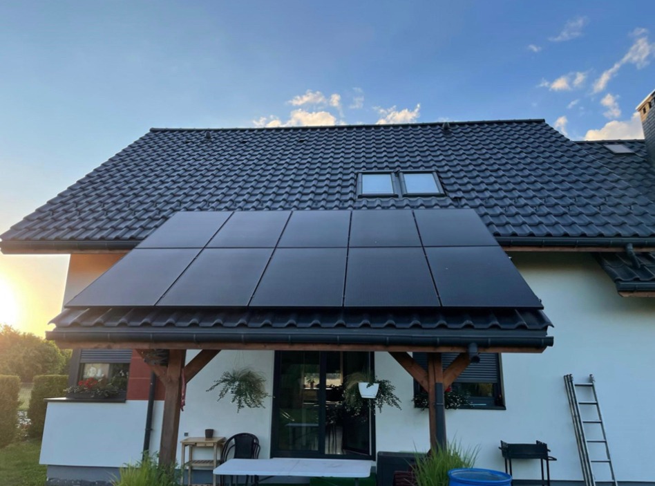 Czarny panel słoneczny zamontowany nad tarasem domu jednorodzinnego z ciemną dachówką, widoczne rośliny w donicach i drabina oparta o ścianę