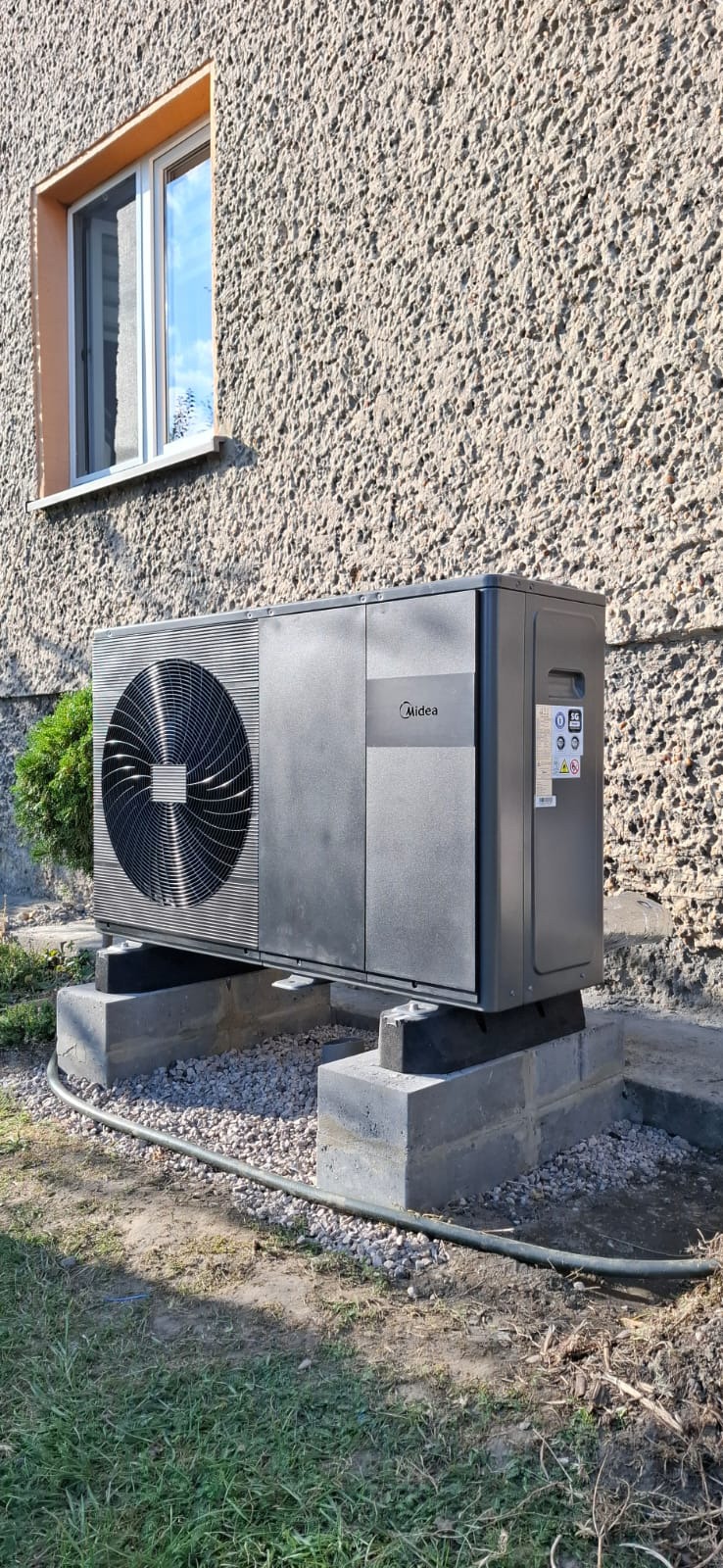 Szara pompa ciepła Midea zamontowana na betonowych blokach fundamentowych przed budynkiem z oknem.