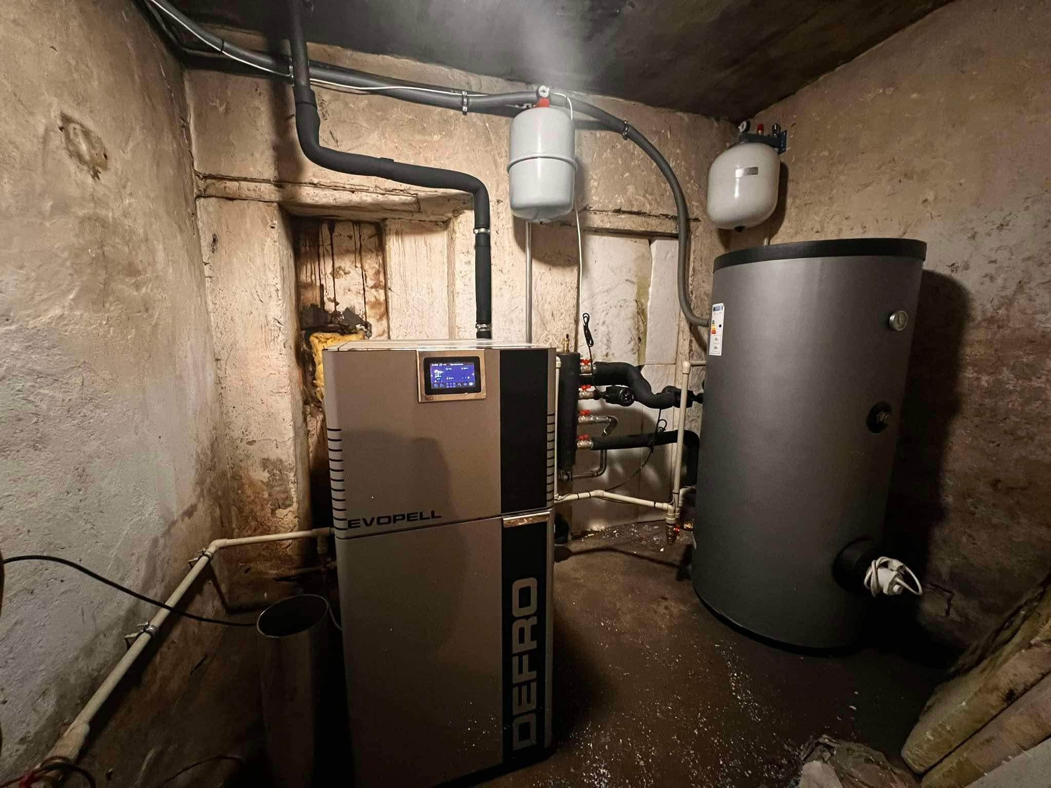Kotłownia z kotłem na pellet marki Defro Evopell i zbiornikiem akumulacyjnym w pomieszczeniu o surowych, betonowych ścianach. Widoczne rury i osprzęt instalacyjny.