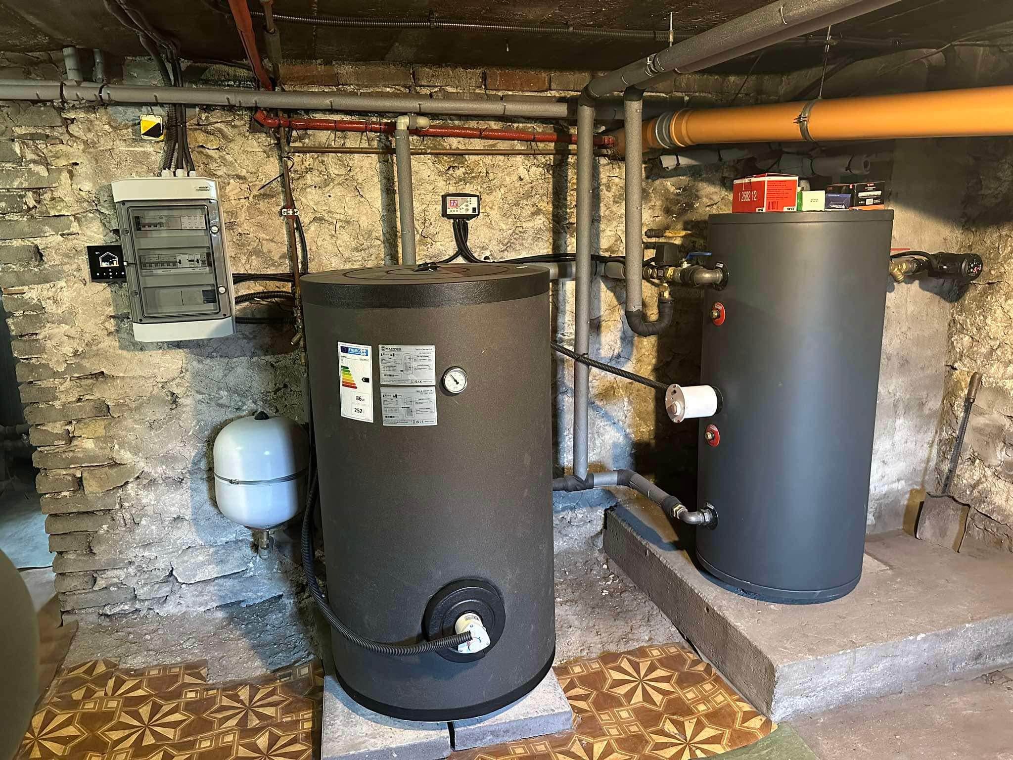 Dwa zbiorniki akumulacyjne w piwnicy z kamiennymi ścianami i widoczną instalacją hydrauliczną, obok skrzynka elektryczna i naczynie przeponowe.