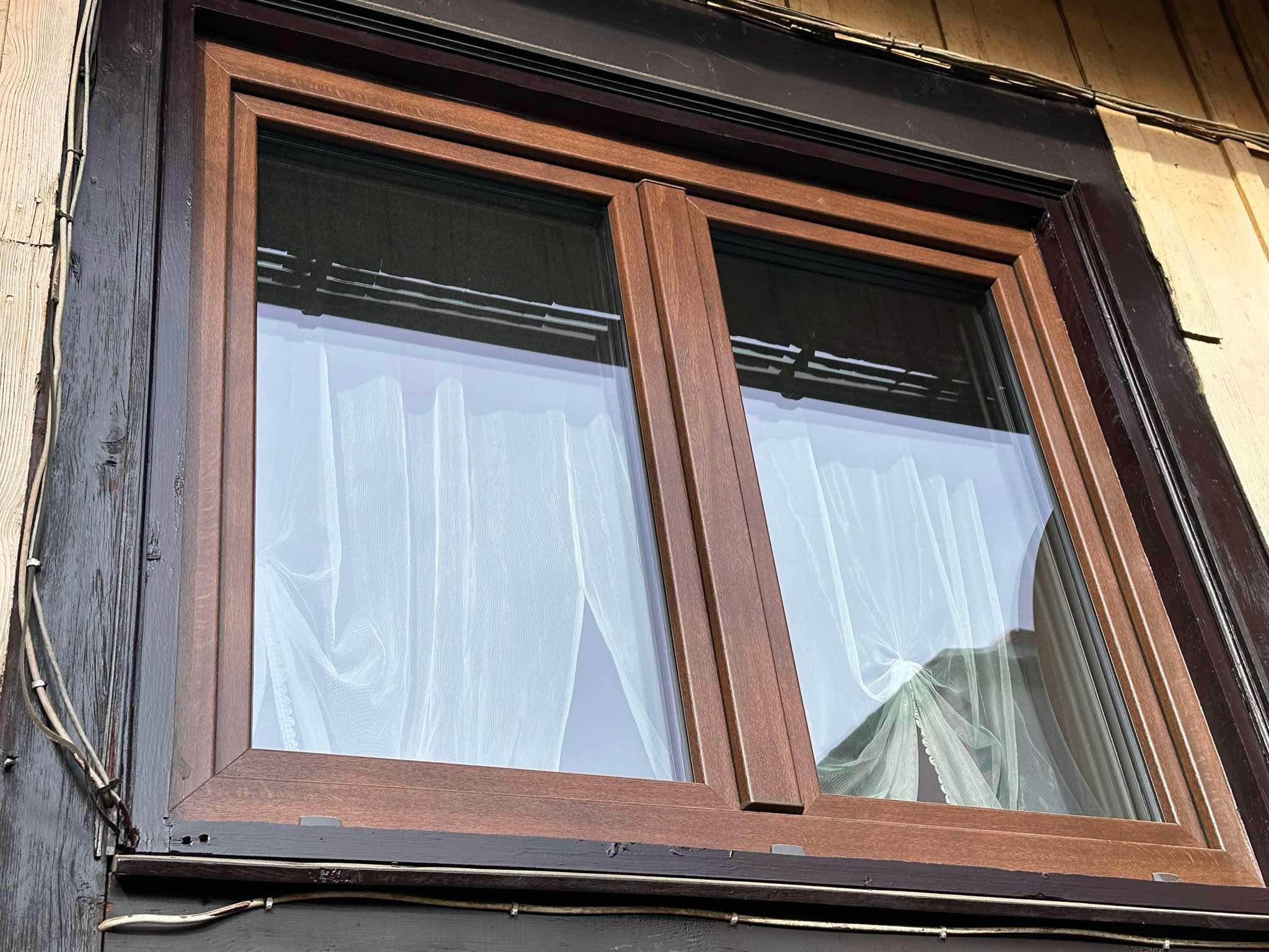 Drewniane okno dwuskrzydłowe z białymi firankami w domu z drewnianą elewacją, widoczne kable na zewnątrz.
