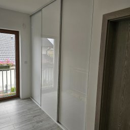 InMeble Bartosz Mielczarek - Biała szafa przesuwna z lustrzanymi drzwiami w jasnym wnętrzu, widok na balkon z kwiatami przez okno.