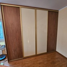 InMeble Bartosz Mielczarek - Przestronna szafa przesuwna z brązowymi drewnopodobnymi bokami i beżowymi panelami środkowymi, z widoczną złotą ramą, w pokoju z drewnianą podłogą i biurkiem przy oknie.