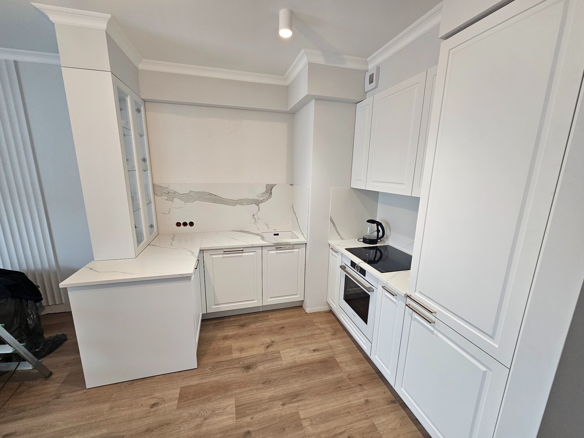 Jasna kuchnia z białymi, klasycznymi szafkami, marmurowym blatem i drewnianą podłogą. Widoczne sprzęty AGD w zabudowie i czajnik na blacie.