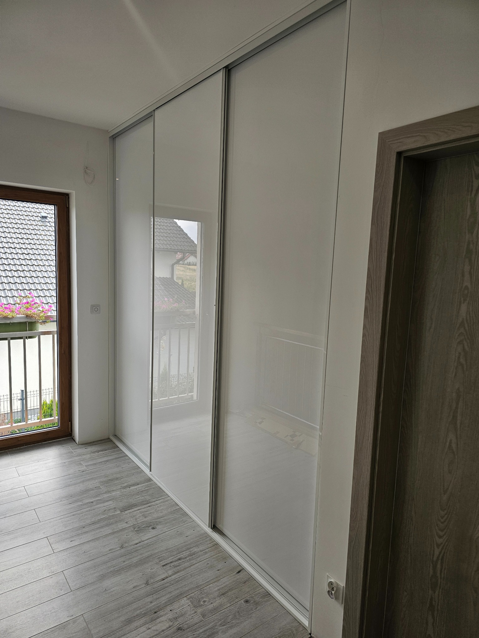 Biała szafa przesuwna z lustrzanymi drzwiami w jasnym wnętrzu, widok na balkon z kwiatami przez okno.