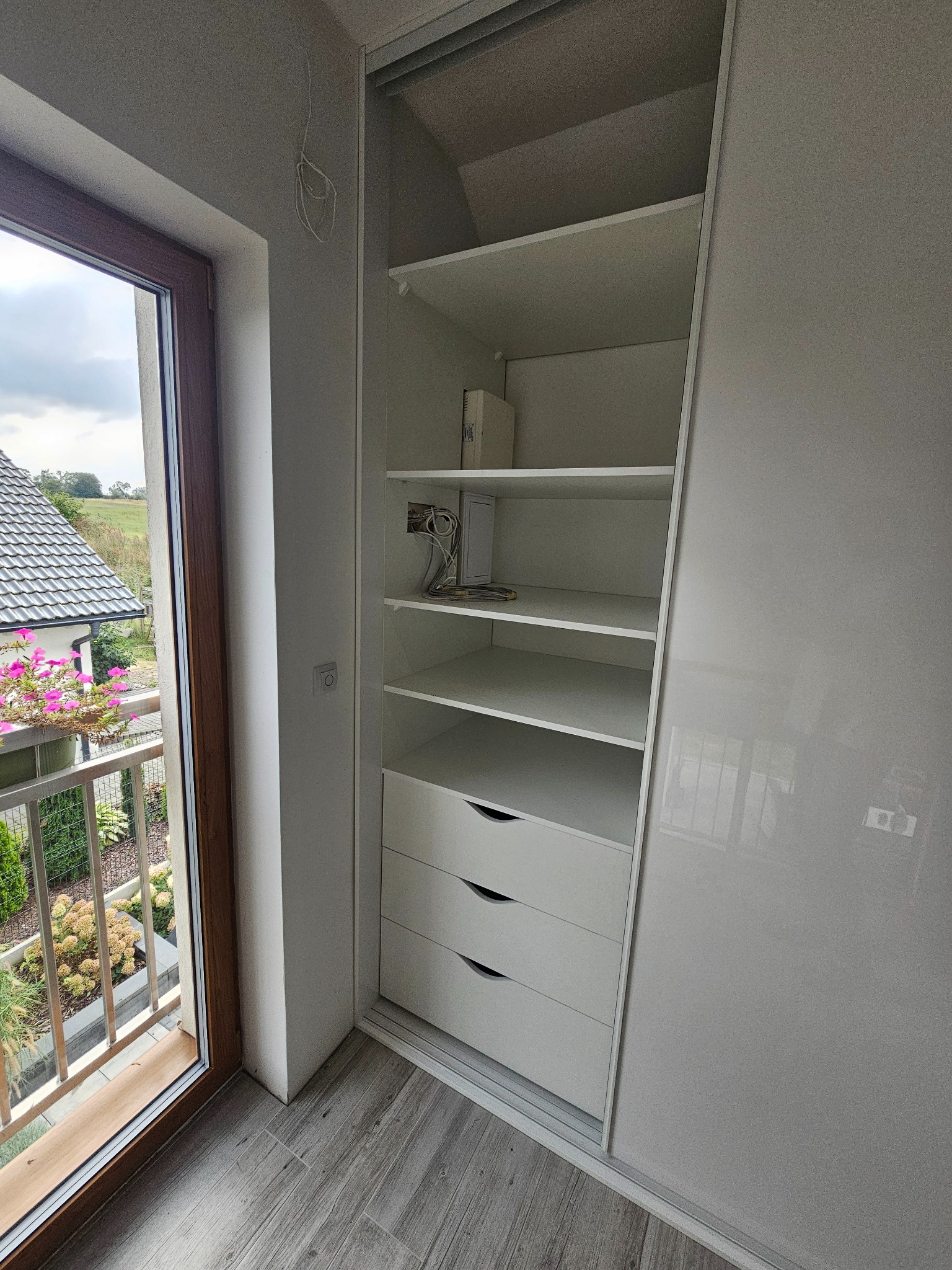 Biała szafa wnękowa z przesuwnymi drzwiami, widoczne półki i trzy szuflady, na ścianie przyłącze elektryczne, z boku okno balkonowe z widokiem na ogród