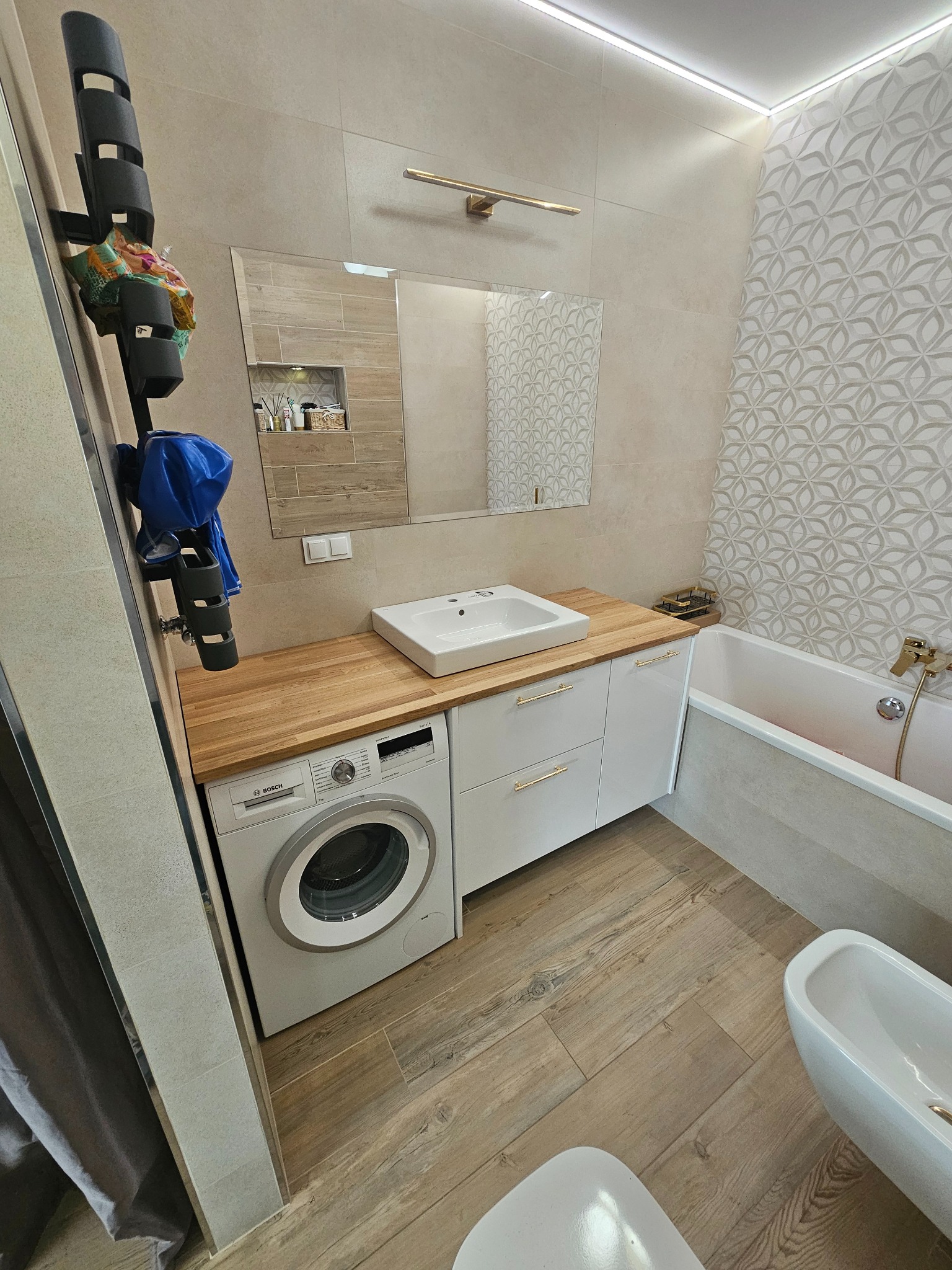 Jasna łazienka z drewnianą podłogą, białymi szafkami pod umywalką z drewnianym blatem, pralką Bosch, wanną i dekoracyjną ścianą w kwiatowy wzór.