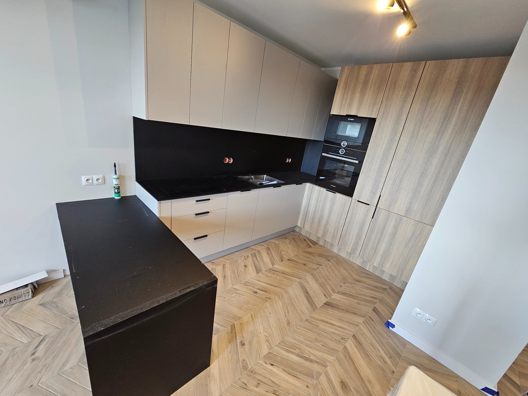 Nowoczesna kuchnia z jasnymi szafkami górnymi i drewnopodobnymi dolnymi, czarny blat roboczy i ściana nad blatem, parkiet ułożony w jodełkę, widoczna tuba z silikonem na blacie.