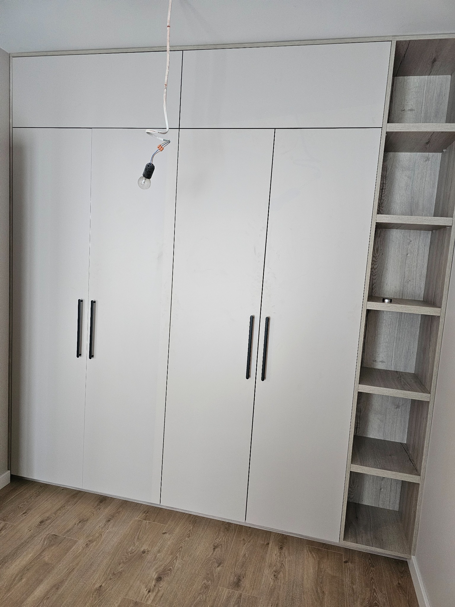Jasnoszara szafa z czterema drzwiami z podłużnymi czarnymi uchwytami i otwartymi półkami z boku, widoczna żarówka na kablu, drewniana podłoga.