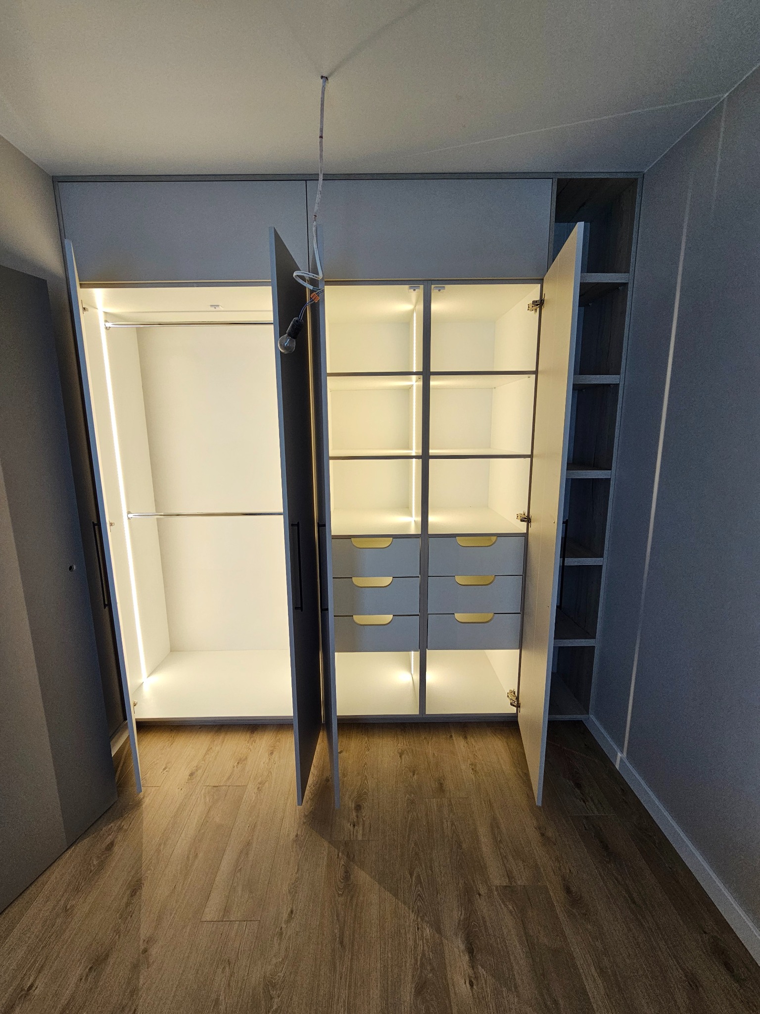 Wnętrze nowoczesnej garderoby z oświetleniem LED, widoczne półki, szuflady z uchwytami, drążki na ubrania, otwarte drzwi, drewniana podłoga, niedokończone podłączenie elektryczne na suficie.