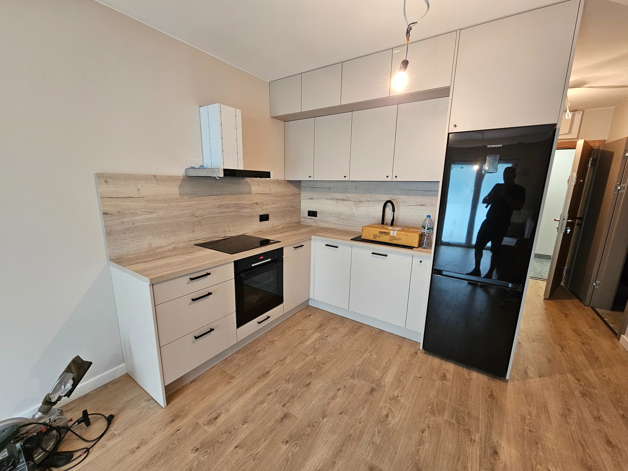 Nowoczesna kuchnia z białymi szafkami i drewnopodobnym blatem, czarna płyta indukcyjna i piekarnik, czarna lodówka Samsung, na podłodze panele imitujące drewno, widoczna lampa wisząca na kablu...