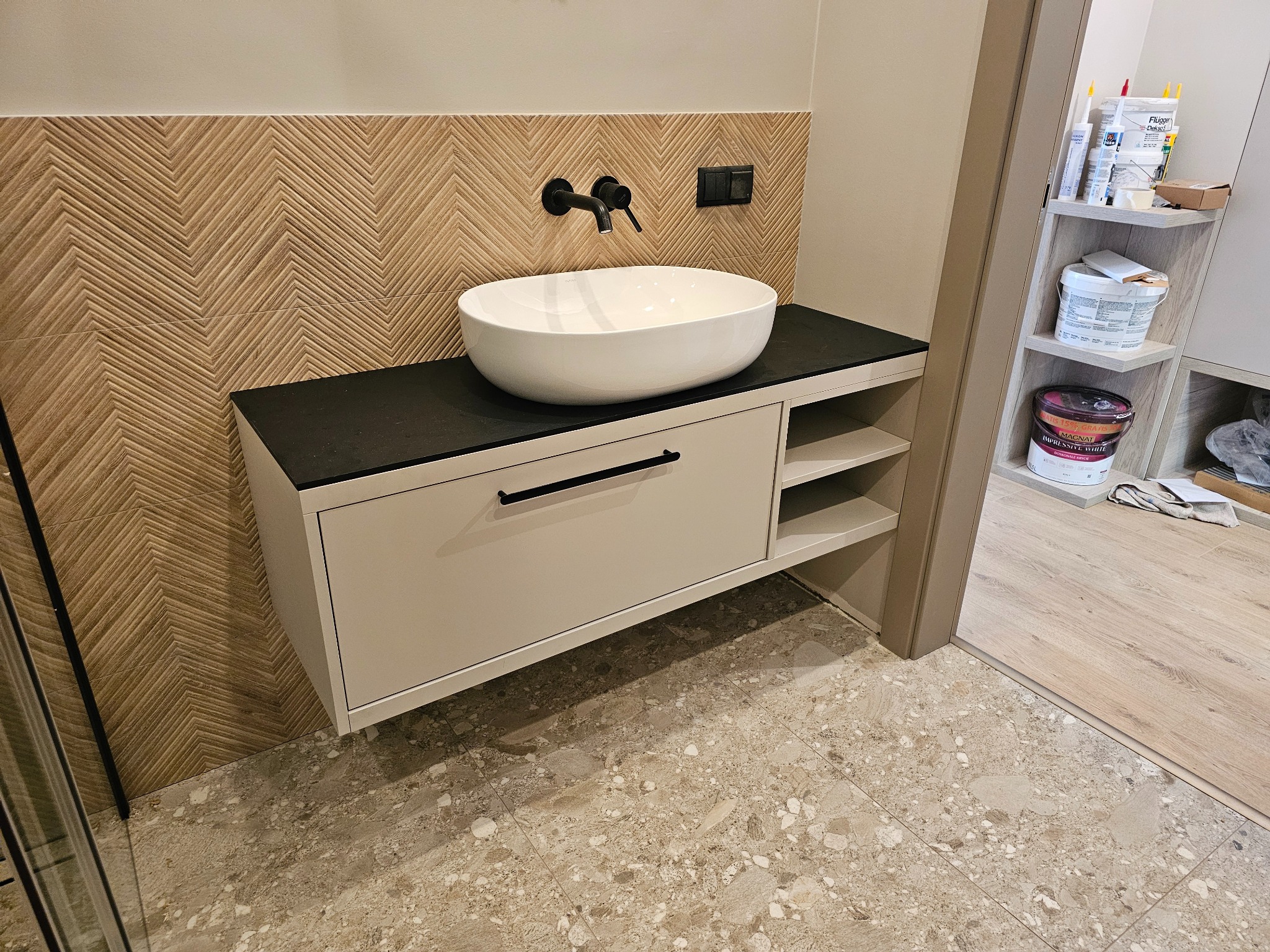 Nowoczesna łazienka z umywalką nablatową i szafką w odcieniach beżu, wzorzysta ściana z płytek imitujących drewno ułożonych w jodełkę, czarna armatura, podłoga z płytek lastryko.