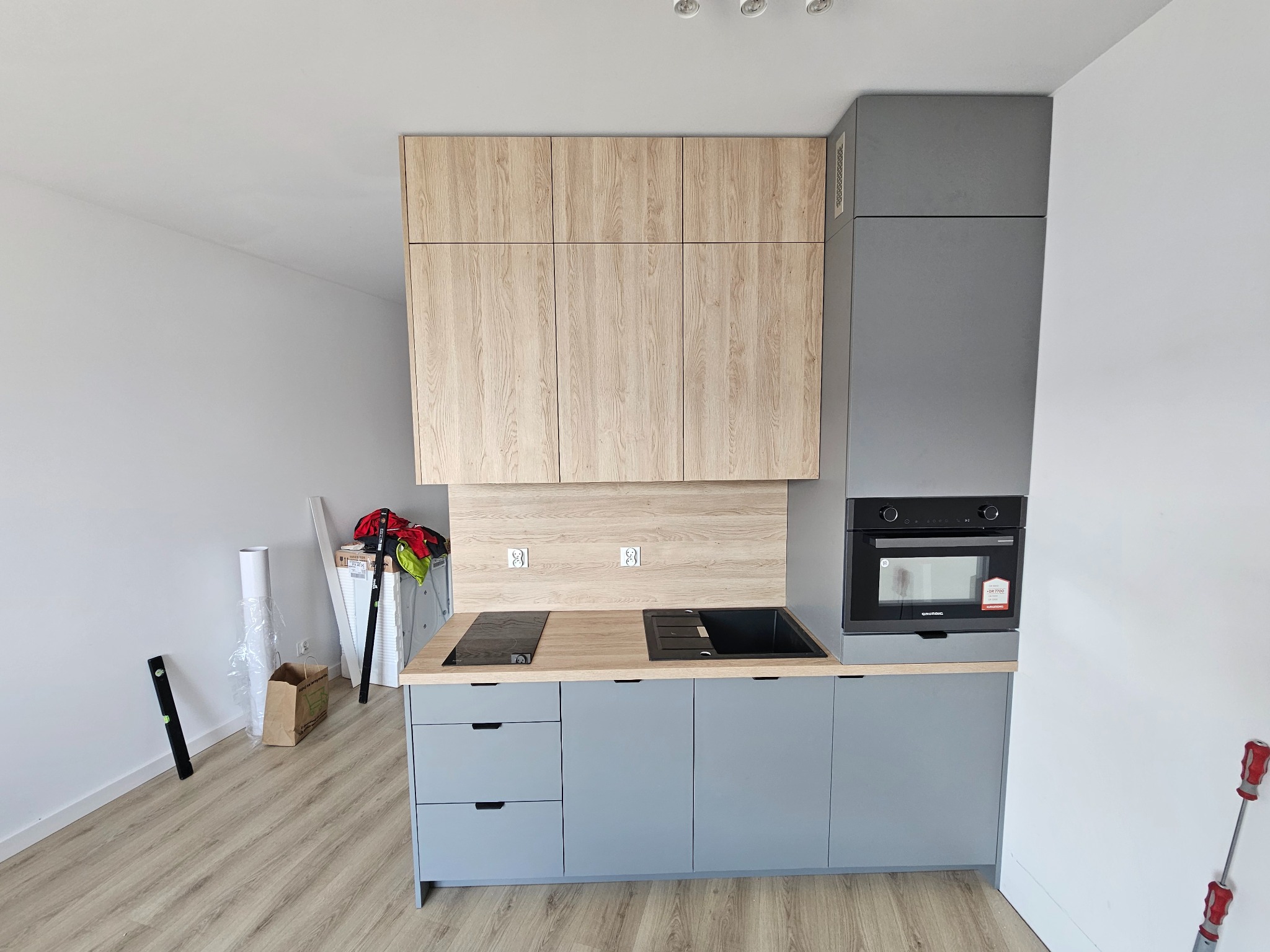 Fragment szarej kuchni z drewnianymi górnymi szafkami i blatem, czarnym zlewem i płytą indukcyjną, widoczne narzędzia i materiały wykończeniowe wskazują na trwający montaż.