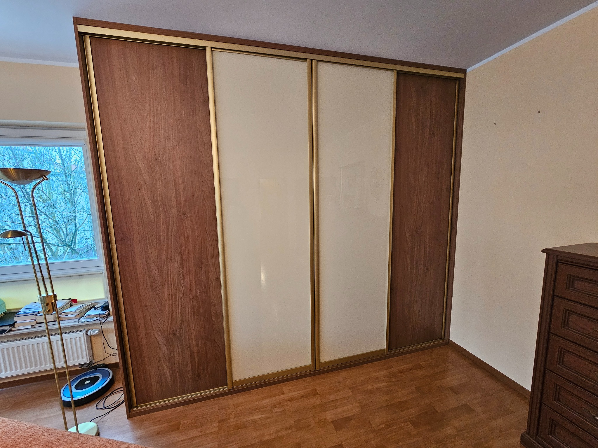 Przestronna szafa przesuwna z brązowymi drewnopodobnymi bokami i beżowymi panelami środkowymi, z widoczną złotą ramą, w pokoju z drewnianą podłogą i biurkiem przy oknie.