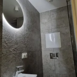 Nowoczesna łazienka z szarą, teksturowaną okładziną ścienną, okrągłym lustrem z podświetleniem LED, białą umywalką narożną i wiszącą toaletą ze srebrnym przyciskiem spłukującym.