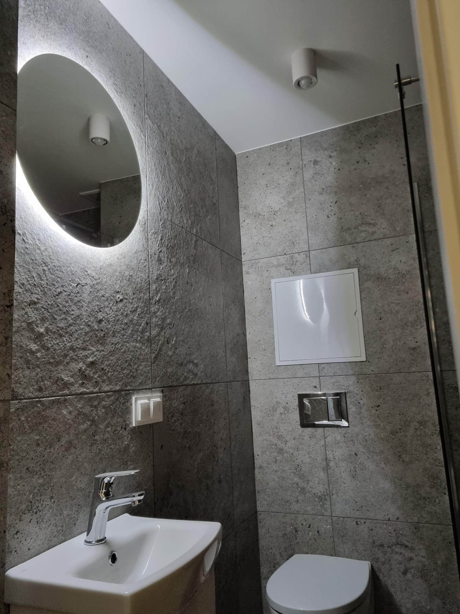 Nowoczesna łazienka z szarą, teksturowaną okładziną ścienną, okrągłym lustrem z podświetleniem LED, białą umywalką narożną i wiszącą toaletą ze srebrnym przyciskiem spłukującym.