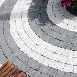 Okrągły wzór z kostki brukowej w odcieniach szarości i bieli, uzupełniony fioletowymi kwiatami i szklanką z kwiatem na drewnianym stoliku.