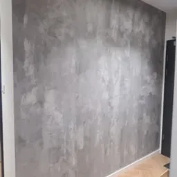 Ściana w pokoju wykończona dekoracyjną szarą fakturą, listwa przypodłogowa w kolorze białym, parkiet ułożony w jodełkę, widoczny fragment gniazdka elektrycznego.