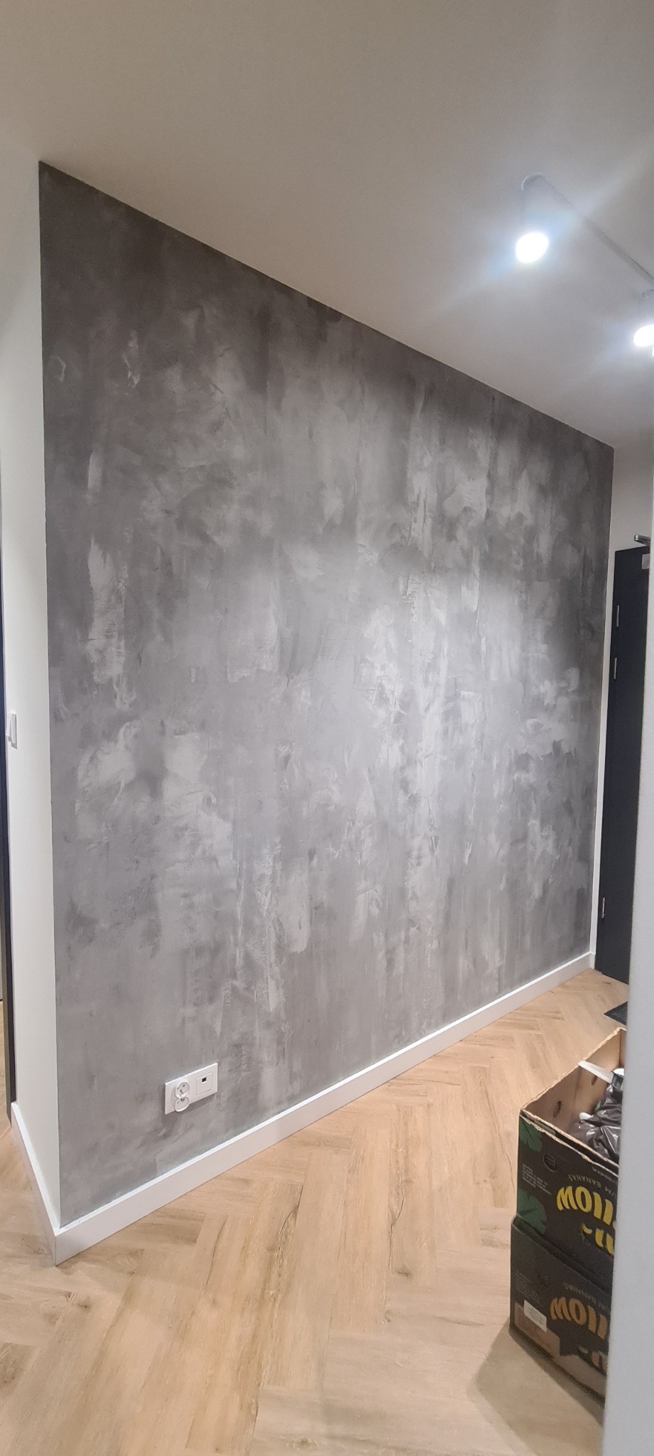 Ściana w pokoju wykończona dekoracyjną szarą fakturą, listwa przypodłogowa w kolorze białym, parkiet ułożony w jodełkę, widoczny fragment gniazdka elektrycznego.