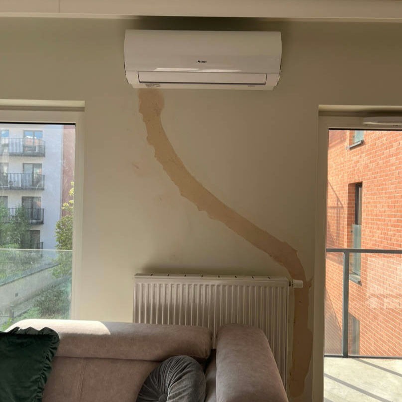 Ściana w pokoju z widocznym śladem po instalacji klimatyzacji, wstępnie pokryta masą wyrównującą, nad grzejnikiem i częściowo zasłonięta przez sofę.