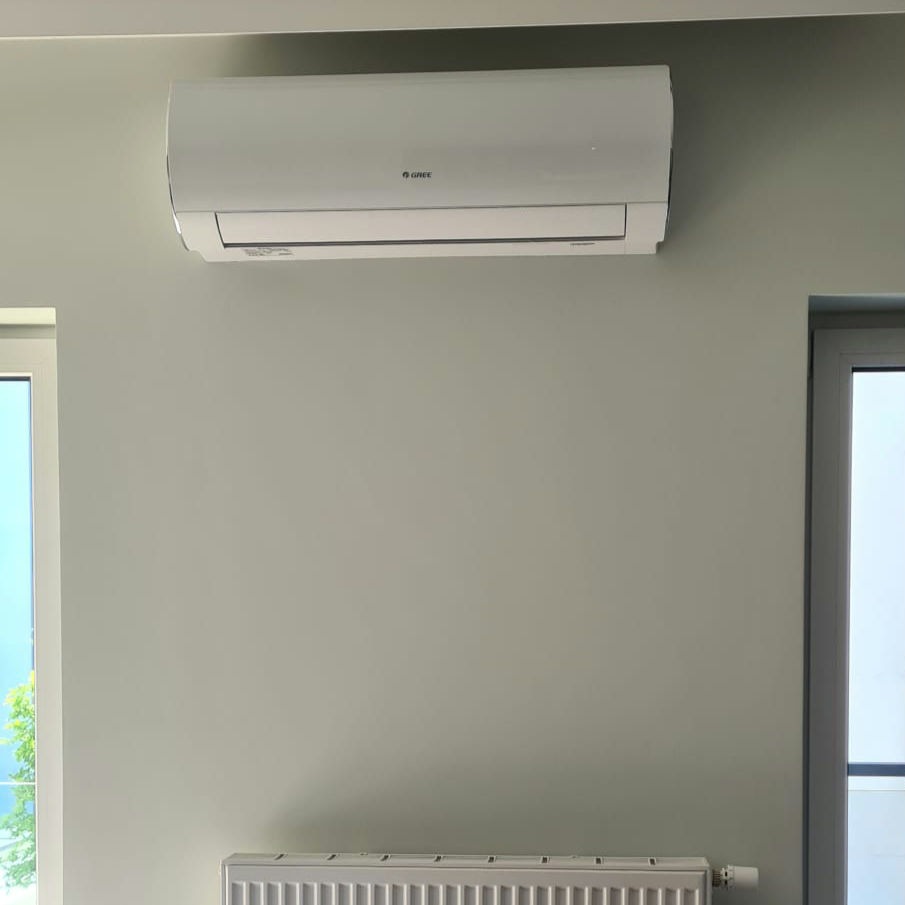 Jasnoszara ściana z zamontowanym klimatyzatorem Gree, widoczne okno i grzejnik.