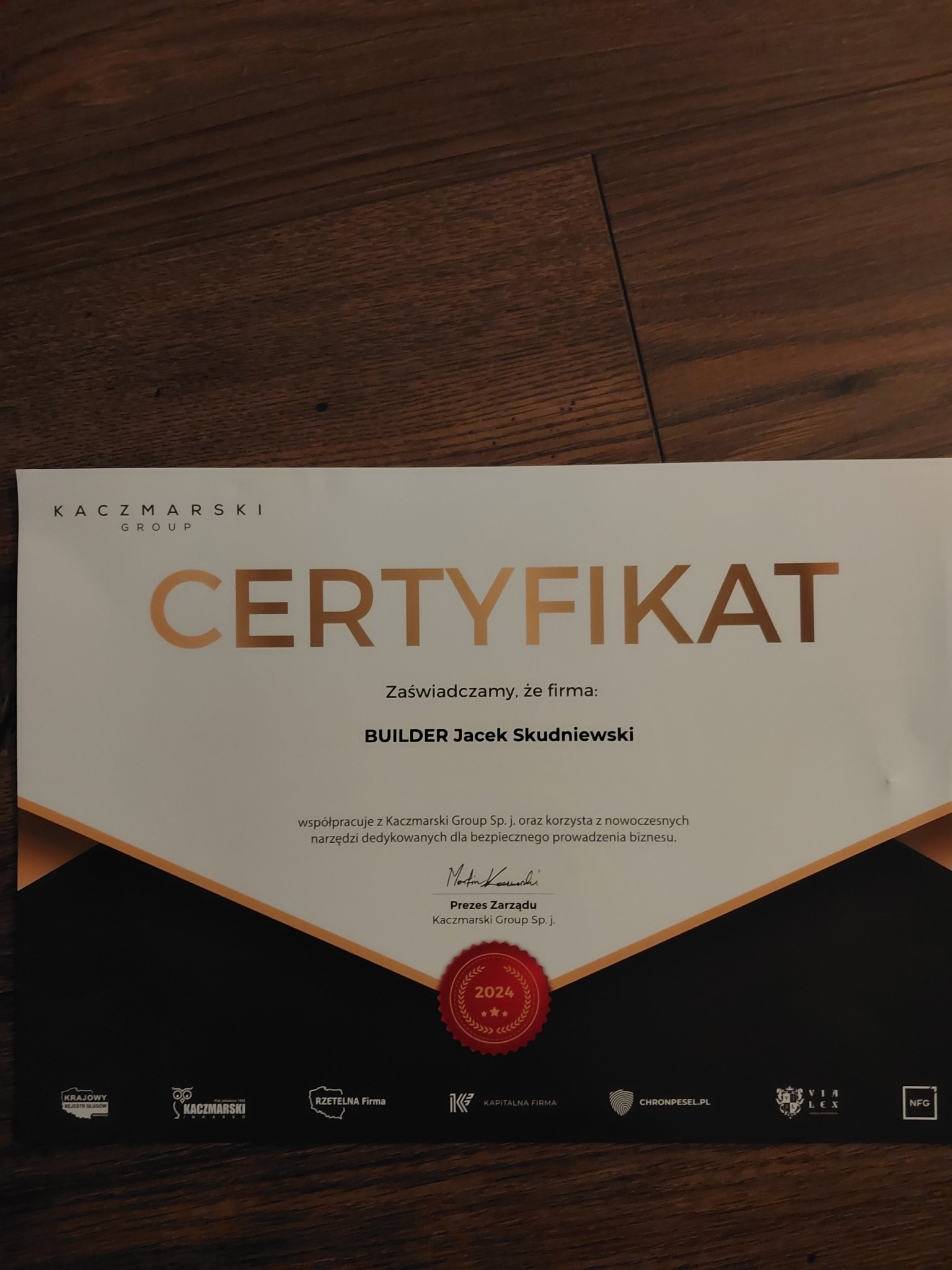 Certyfikat firmy Builder Jacek Skudniewski na drewnianym tle, potwierdzający współpracę z Kaczmarski Group.