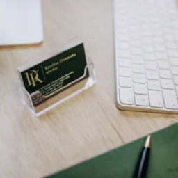 Wizytówki adwokackie w przezroczystym stojaku, obok klawiatura Apple, notatnik i długopis na drewnianym biurku. Widoczne logo kancelarii i dane kontaktowe adwokata.