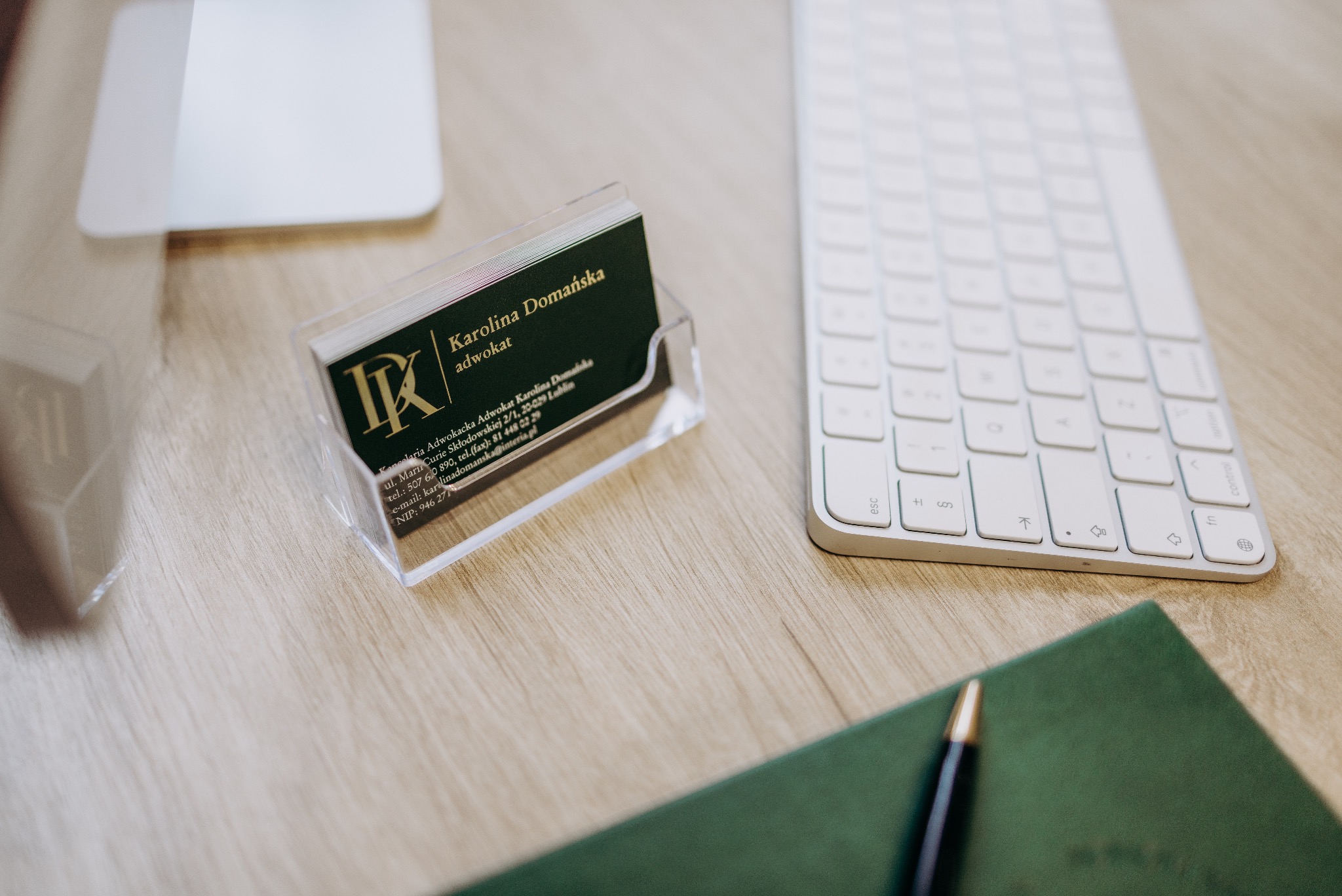 Wizytówki adwokackie w przezroczystym stojaku, obok klawiatura Apple, notatnik i długopis na drewnianym biurku. Widoczne logo kancelarii i dane kontaktowe adwokata.