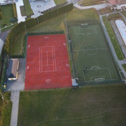 Ordofly - Widok z lotu ptaka na kompleks boisk sportowych: czerwone boisko do koszykówki i zielone boisko do piłki nożnej, obok plac zabaw dla dzieci, otoczone zielenią i zabudową mieszkalną.