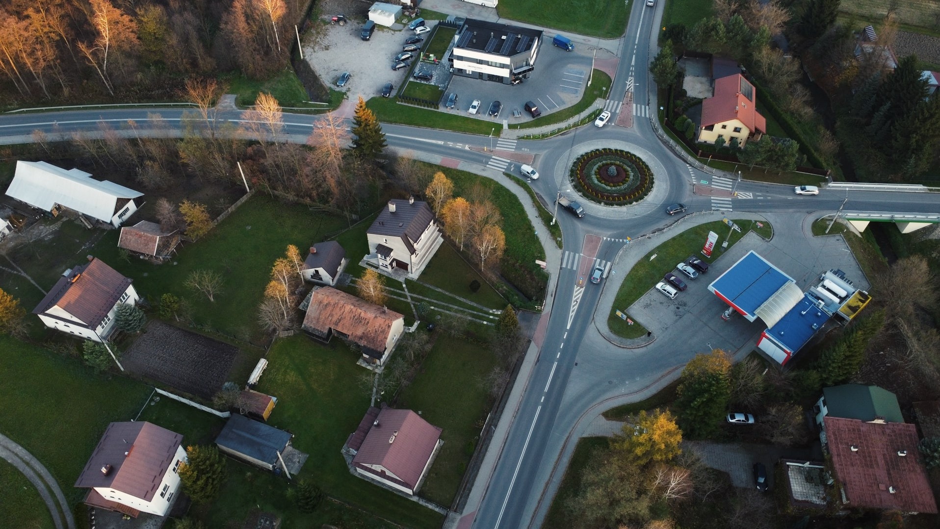 Zdjęcie z drona przedstawia skrzyżowanie z rondem, budynki mieszkalne, stację benzynową i budynek biurowy z parkingiem, otoczone drzewami w jesiennej scenerii.