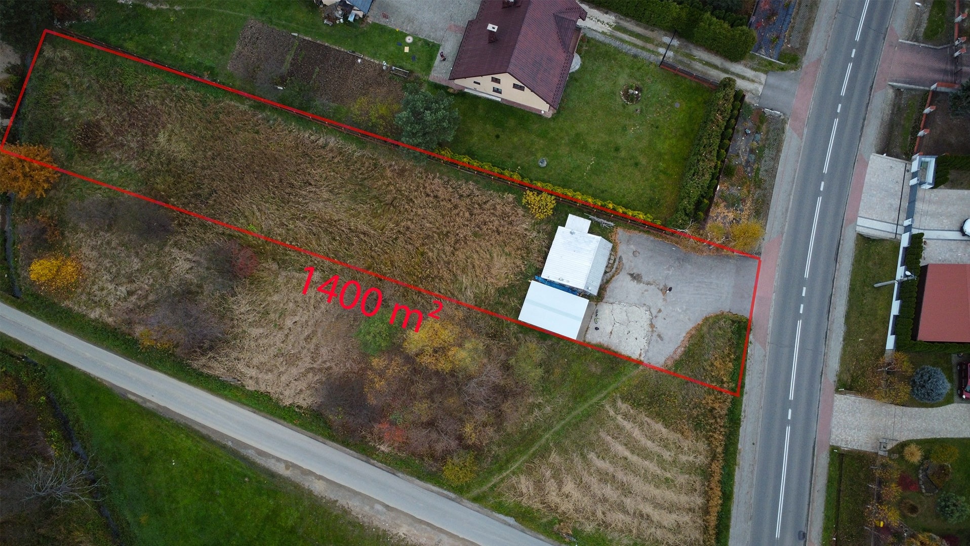 Zdjęcie z lotu ptaka przedstawiające działkę o powierzchni 1400 m² zarysowaną czerwoną linią, sąsiadującą z drogą i zabudowaniami.