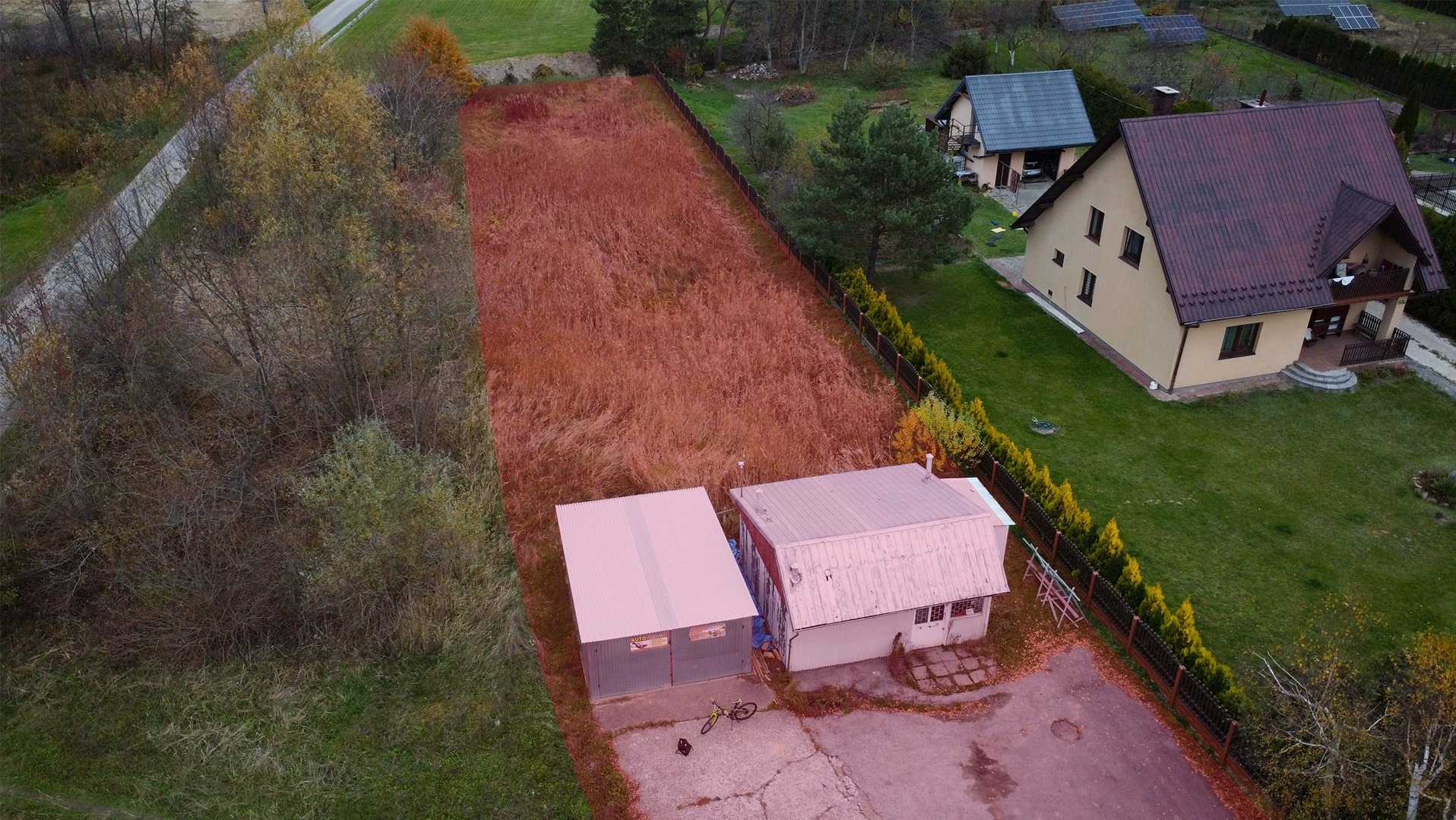 Widok z drona na działkę z budynkami gospodarczymi, zarośniętą trawą w kolorze rdzy, obok dom z brązowym dachem i panele słoneczne w tle.