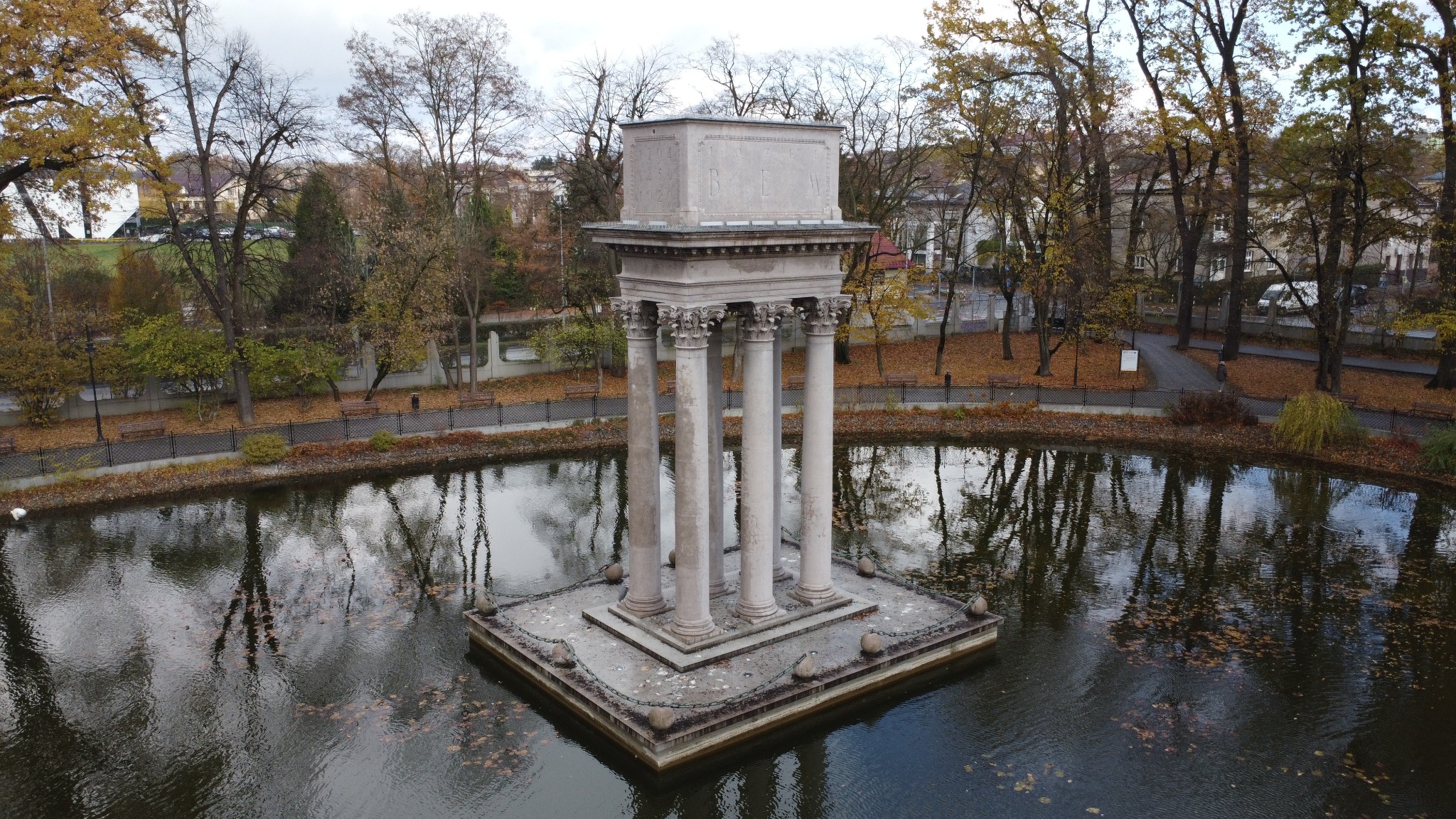 Klasycystyczna budowla z kolumnami na platformie otoczonej wodą w jesiennym parku miejskim, widok z góry.